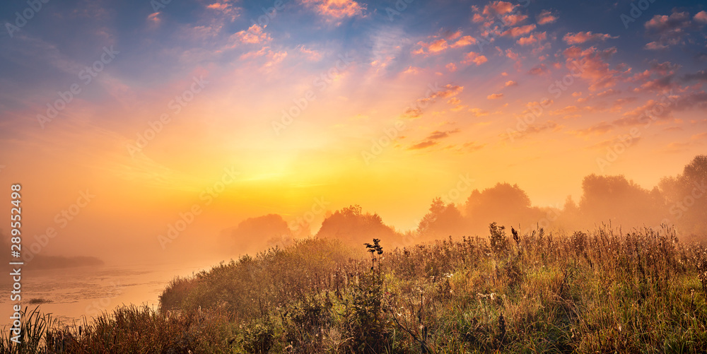 Beautiful golden sunrise panorama at the misty riverside with the bright sun shining through the clouds