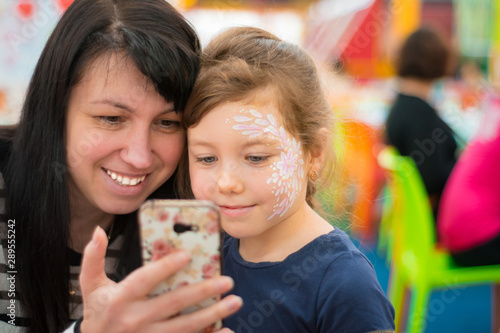 Mom and daughter are looking at a mobile phone. Girls make selfie in a cafe catering. A child in a fashionable makeup with his mother using the phone.