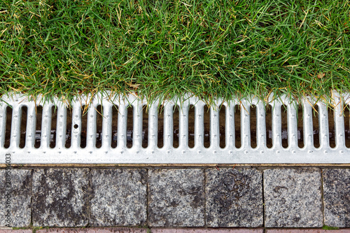 iron grate of a storm drainage system on the side of a footpath made of pavers near a green lawn top view. photo