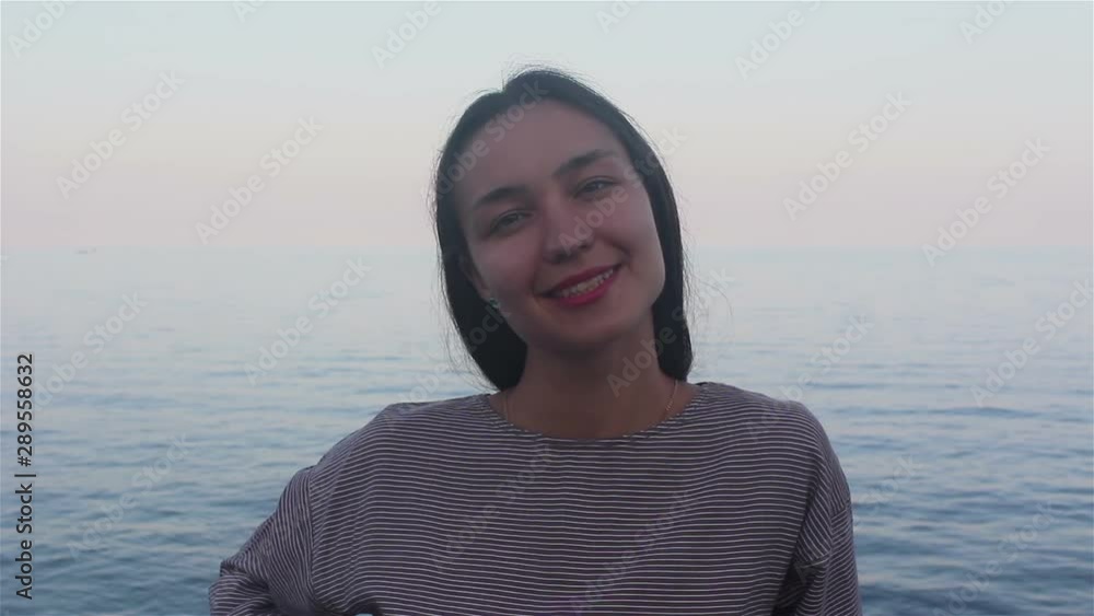 Portrait of a young beautiful girl on the background of the blue sea.Close-up portrait of an attractive girl on the background of the sea and beaches, she looks at the camera, smiles and waves her han Portrait of a young beautiful girl on the background of the blue sea.Close-up portrait of an attractive girl on the background of the sea and beaches, she looks at the camera, smiles and waves her han