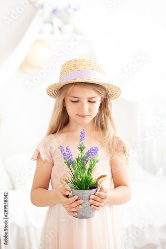 Cute little girl with long blond hair in a straw hat in a dress holds pot of lavender. Childhood concept. Gardening. floriculture. Environmental Protection. Hobby. Summer flowers, houseplant lavender