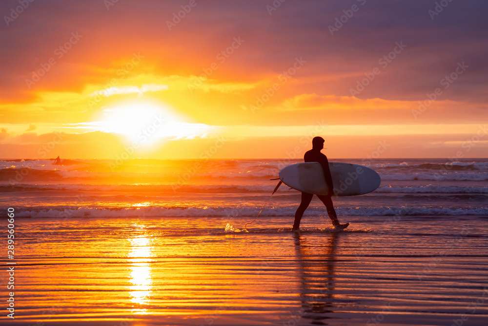 Surfer walking towards the Ocean on the sandy beach during a dramatic summer sunset. Taken in Seaside, Oregon, United States of America.