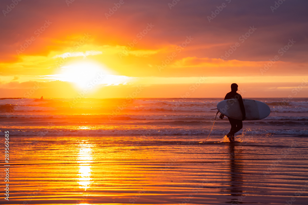 Surfer walking towards the Ocean on the sandy beach during a dramatic summer sunset. Taken in Seaside, Oregon, United States of America.