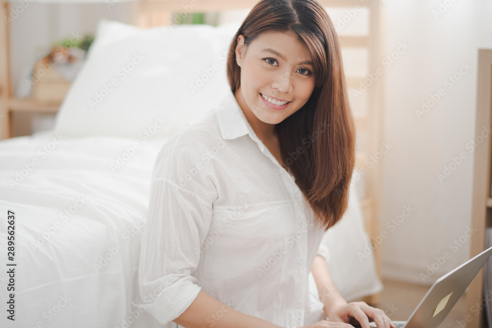Young beautiful Asian woman sitting on ground floor near white bed and working by using computer laptop looking at camera with glad smiling and felling happy. work at home for woman life style Young beautiful Asian woman sitting on ground floor near white bed and working by using computer laptop looking at camera with glad smiling and felling happy. work at home for woman life style