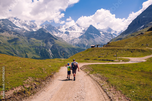 Grandfather and grandson enjoy a great view while hiking a mountain trail high in the Pennine Alps in summer, rear view. Valais, Switzerland
