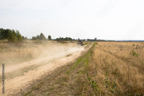 Caucasian man in sport protective costume riding an ATV quad bike over rough terrain with meadows of dry autumn grass. Adventure. Road to the field, dust from under the wheels. © evgeniusd