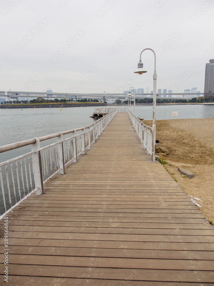 Wide vertical view of an old wooden pier with steel railings along the oceanfront bay on a cloudy day. Odaiba Island, Tokyo, Japan