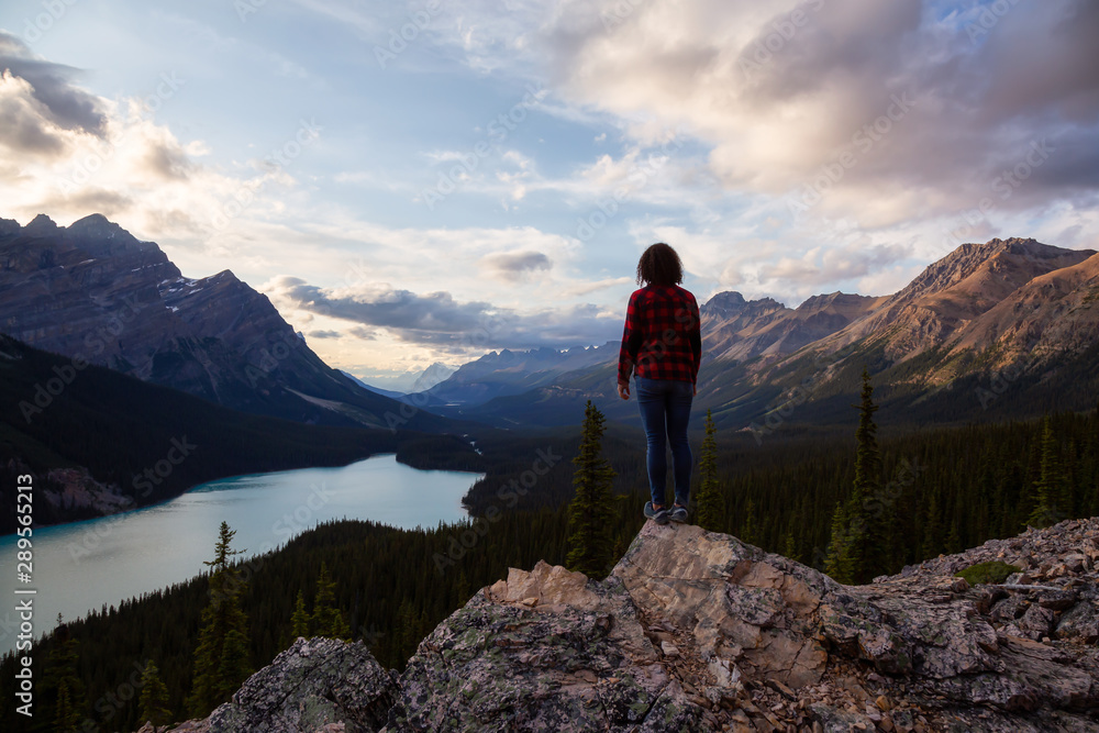 Fototapeta premium Adventurous girl standing on the edge of a cliff overlooking the beautiful Canadian Rockies and Peyto Lake during a vibrant summer sunset. Taken in Banff National Park, Alberta, Canada.