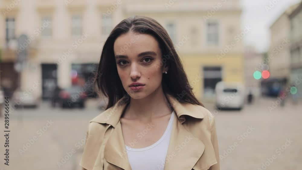 Portrait of Pretty Young Girl Confident Serious Girl Open Eyes Wearing Beige Trench Standing in the Quiet City Street Close Up. Portrait of Pretty Young Girl Confident Serious Girl Open Eyes Wearing Beige Trench Standing in the Quiet City Street Close Up.