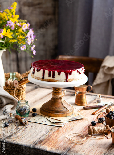Fototapeta Naklejka Na Ścianę i Meble -  homemade tasty whole cheesecake decorated with purple sauce on top served on wooden cake stand on grey table with flowers and berries, selective focus