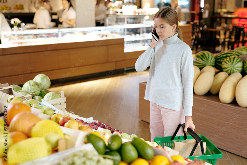 Pretty girl in casualwear talking to her mom on mobile phone by fruit display