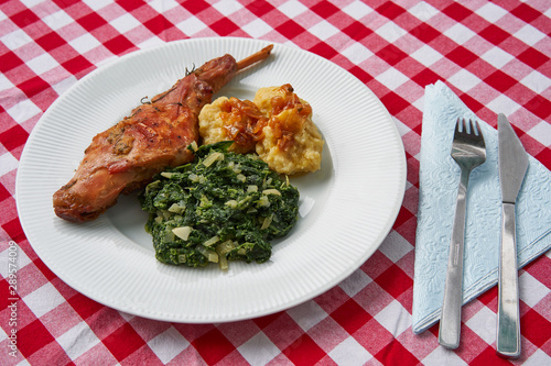 Roasted rabbit leg, marinated in garlic and rosemary served with spinach, chopped onion and potato dumplings on white plate with fork, knife and blue napkin.Ttraditional dish cuisine of Czech republic