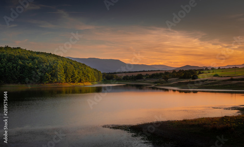 The calm and quiet waters of a mountain lake surrounded by green hills, fields, and forests, distanty mountain range in the background, blue sky with smooth clouds colored in orange by the sunset