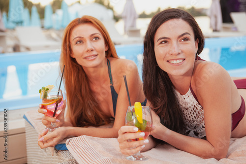 Lovely young women smiling cheerfully, sunbathing near the swimming pool together. Happy beautiful female friends tanning at the poolside. Travel, friendship concept