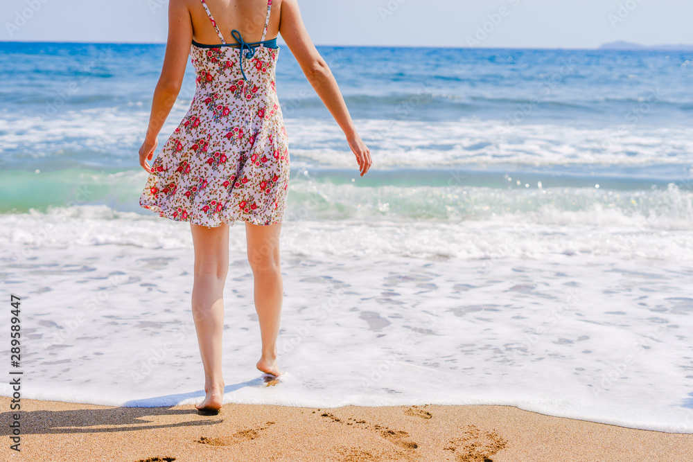 Woman walking on the beach to the sea in summer dress barefoot legs feet with footprints on the vacation holiday beach travel seashore ocean