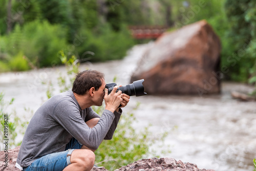 Man taking pictures with camera in Redstone, Colorado during summer with large boulder and red bridge on Crystal river by trees