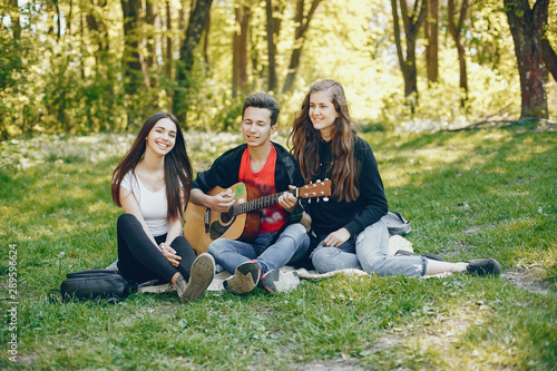Three friends sitting on a green grass. Boy playing on a guitar with his two girlfriends. Two girls have fun with their friend
