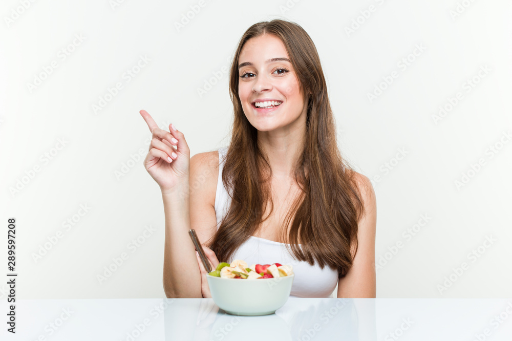 Young caucasian woman eating fruit bowl smiling cheerfully pointing with forefinger away. Young caucasian woman eating fruit bowl smiling cheerfully pointing with forefinger away.