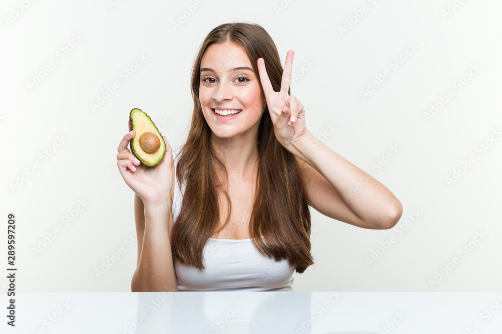 Young caucasian woman holding an avocado showing victory sign and smiling broadly. Young caucasian woman holding an avocado showing victory sign and smiling broadly.