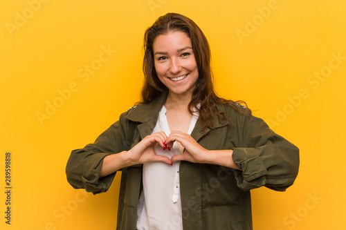 Young european woman isolated over yellow background smiling and showing a heart shape with her hands. © Asier
