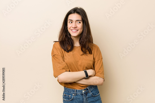 Young brunette woman against a beige background smiling confident with crossed arms.