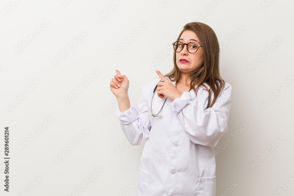 Young doctor woman against a white wall shocked pointing with index fingers to a copy space. Young doctor woman against a white wall shocked pointing with index fingers to a copy space.
