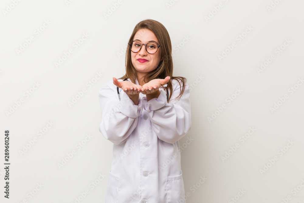 Young doctor woman against a white wall holding something with palms, offering to camera. Young doctor woman against a white wall holding something with palms, offering to camera.