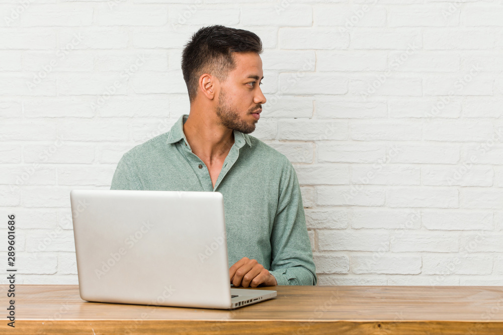 Young filipino man sitting working with his laptop gazing left, sideways pose. Young filipino man sitting working with his laptop gazing left, sideways pose.