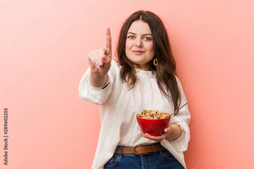 Young plus size curvy woman holding a cereals bowl showing number one with finger. Young plus size curvy woman holding a cereals bowl showing number one with finger.