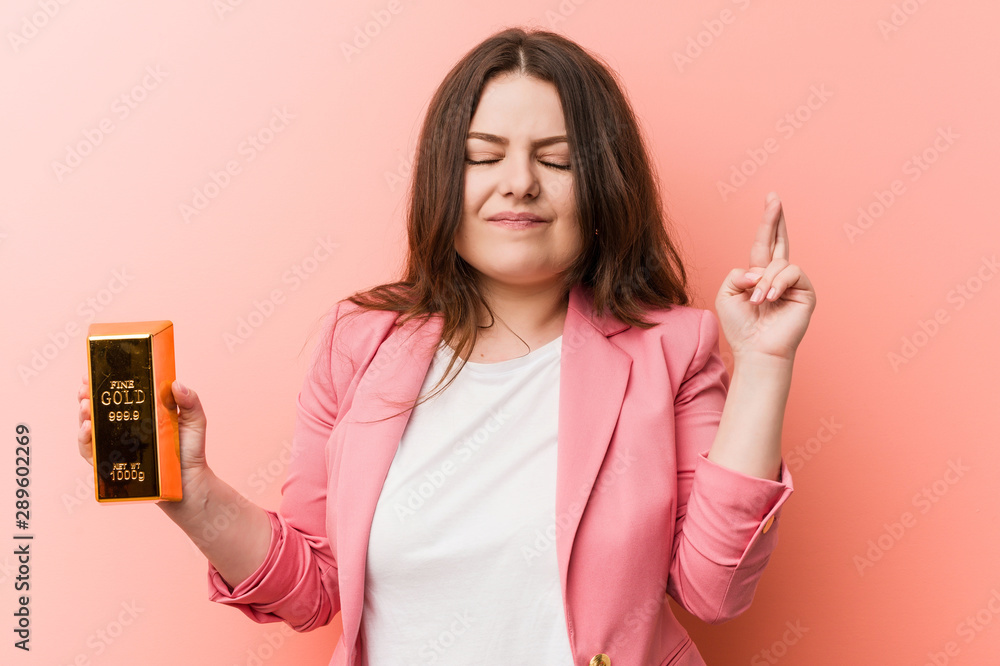 Young plus size curvy business woman holding a gold ingot crossing fingers for having luck Young plus size curvy business woman holding a gold ingot crossing fingers for having luck