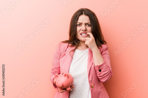 Young plus size curvy business woman holding a piggy bank biting fingernails, nervous and very anxious.