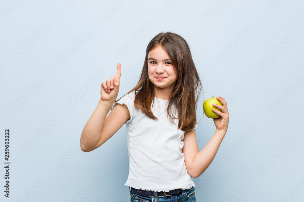 Little caucasian girl holding a green apple showing number one with finger. Little caucasian girl holding a green apple showing number one with finger.