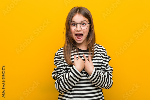 Little caucasian cute girl surprised pointing at himself, smiling broadly.