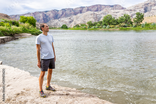 Beach shore view with man standing looking at Green River Camground in Dinosaur National Monument Park with green plants and sand by water © Kristina Blokhin
