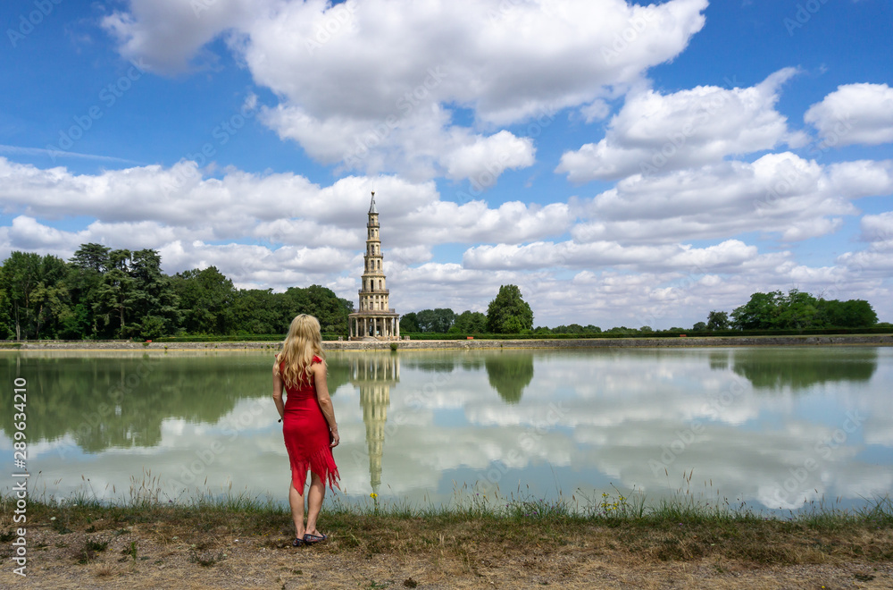 Fototapeta premium Beautiful blonde woman in red dress staring out across an emerald lake toward 18th-century Chanteloup Pagoda.