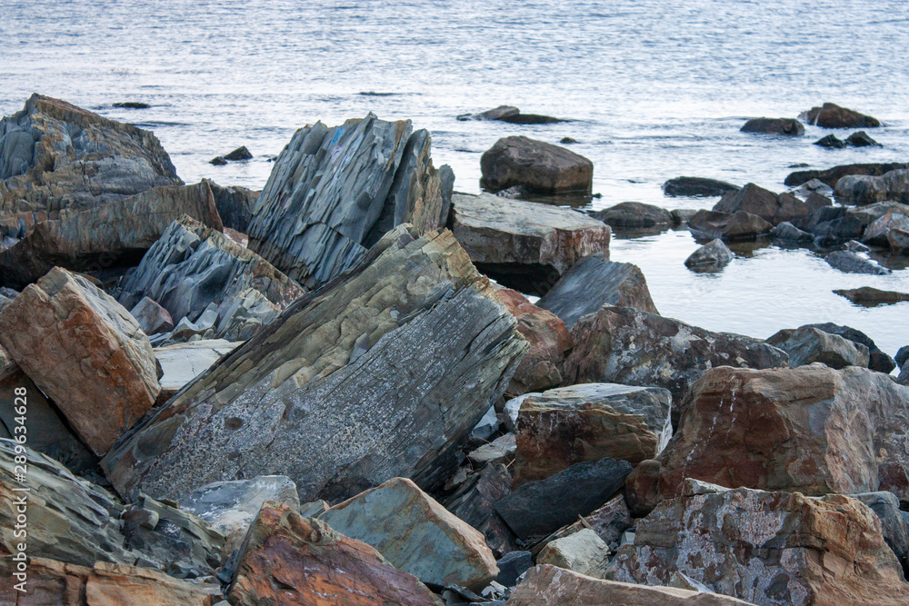 Large fragments of rocks and stones on the seashore. Brown cliffs near calm sea water.