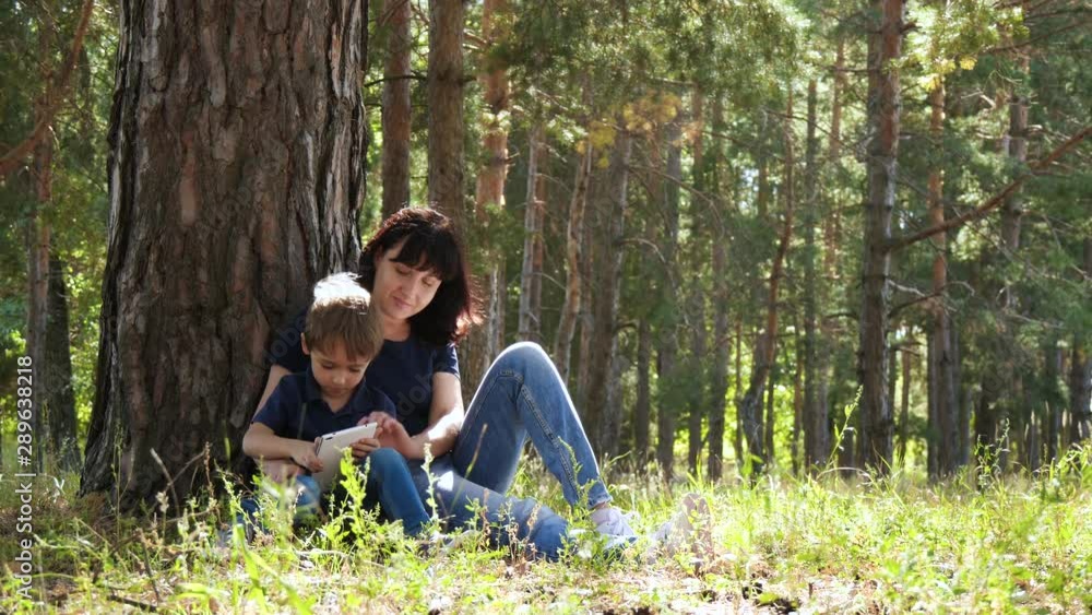 A young mother and her son are relaxing in the Park, sitting by a tree and using a gadget. Contemporary technology. A young mother and her son are relaxing in the Park, sitting by a tree and using a gadget. Contemporary technology.