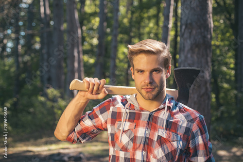 Lumberjack carries axe on shoulder. Deforestation. Handsome man with axe. Lumberjack in the woods with an ax.