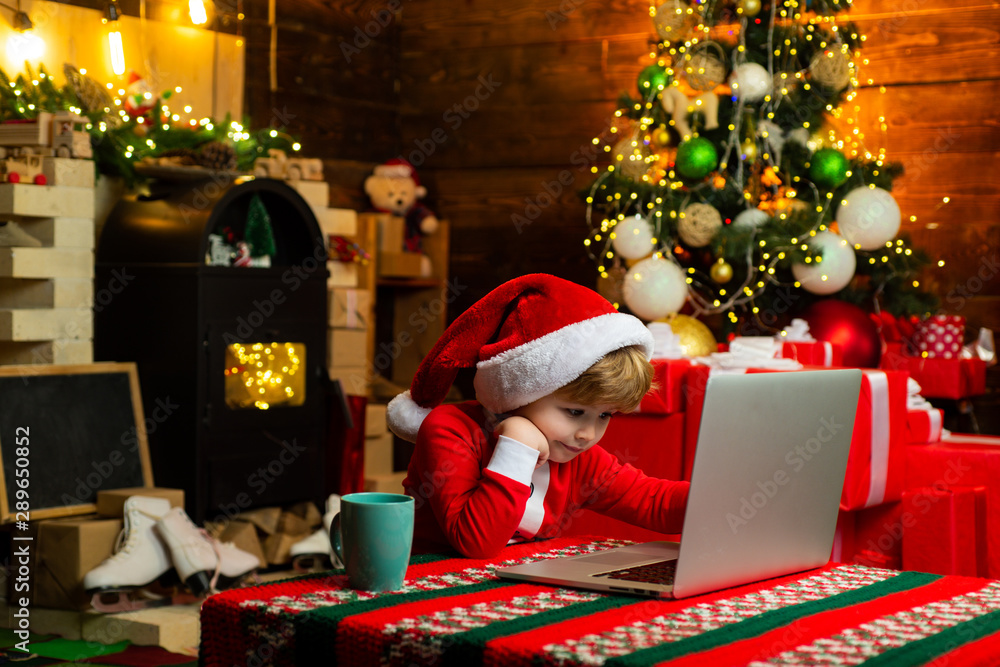 Gifts service. Little genius. Santa little helper. Little boy santa hat and  costume having fun. Boy child with laptop near christmas tree. Buy  christmas gifts online. Christmas shopping concept Stock Photo