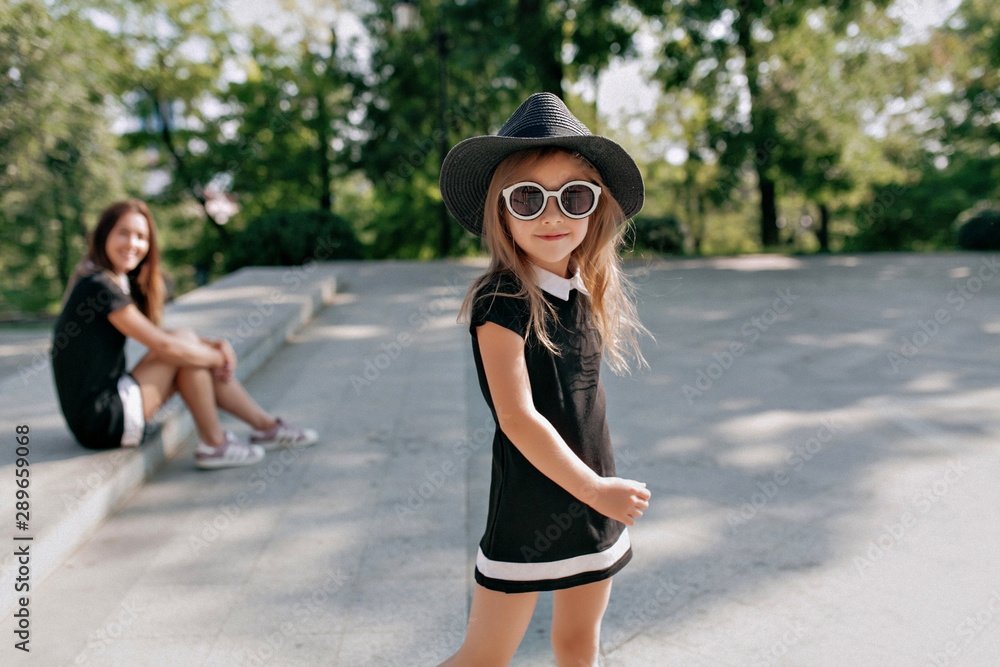 Outsdie portrait of pretty charming young lady wearing black glasses and hat and dress walking with mother in sunny day