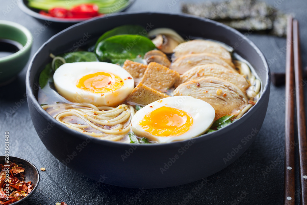 Asian noodle soup, ramen with chicken, tofu, vegetables and egg in black bowl. Slate background. Close up. Asian noodle soup, ramen with chicken, tofu, vegetables and egg in black bowl. Slate background. Close up.