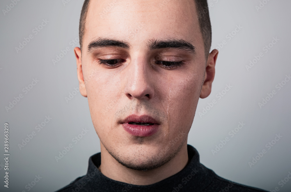 Fototapeta premium Unhappy crying young man with dropping tears on the face. Closeup studio portrait on grey background. Negative emotional concept