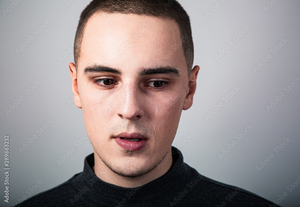 Fototapeta premium Unhappy crying young man with dropping tears on the face. Closeup studio portrait on grey background. Negative emotional concept