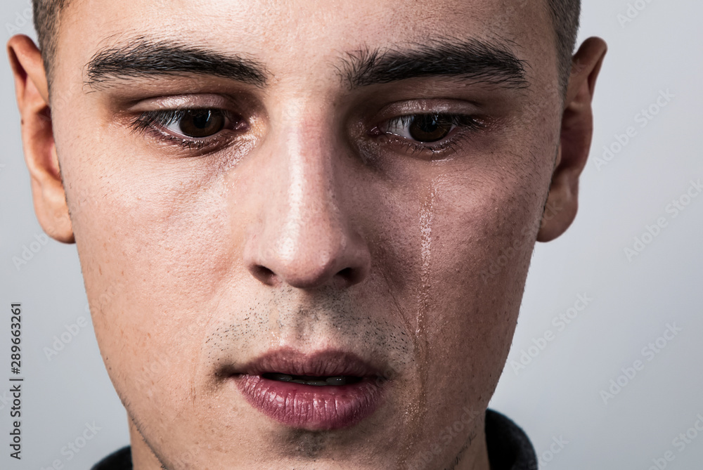 Obraz premium Unhappy crying young man with dropping tears on the face. Closeup studio portrait on grey background. Negative concept