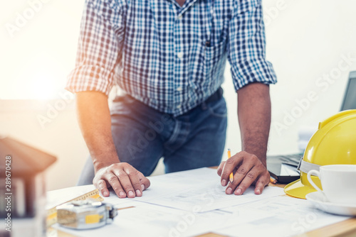 Asian architect man Standing working with blueprints sketching a construction project on wood desk at home office.Construction design concept.