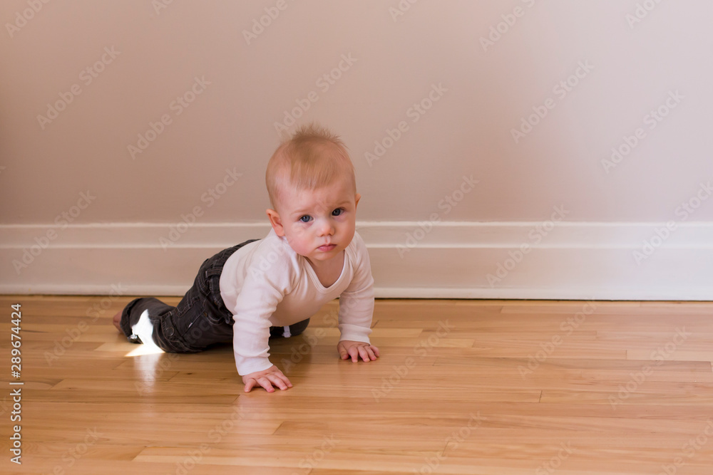 Horizontal photo of adorable barefoot baby boy in casual clothes learning to crawl on hardwood floor looking up with wary expression