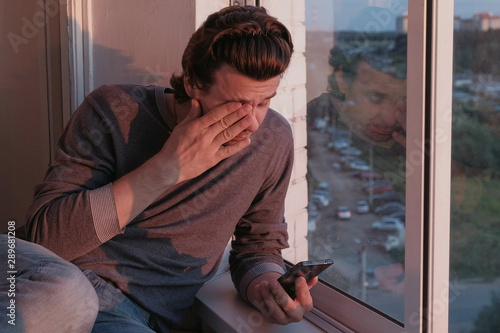 Tired man looks at mobile phone, flipping the tape in social networks sitting on balcony with city view at sunset in evening © Evgeniia