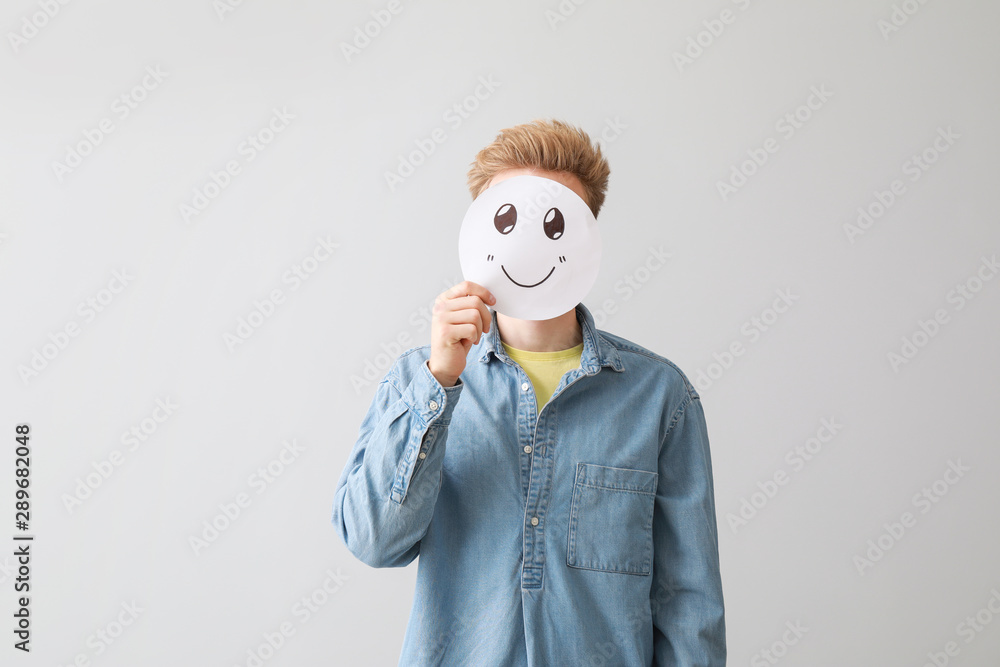 Man hiding face behind emoticon on light background