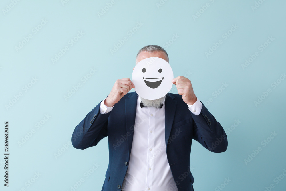 Mature man hiding face behind emoticon on color background