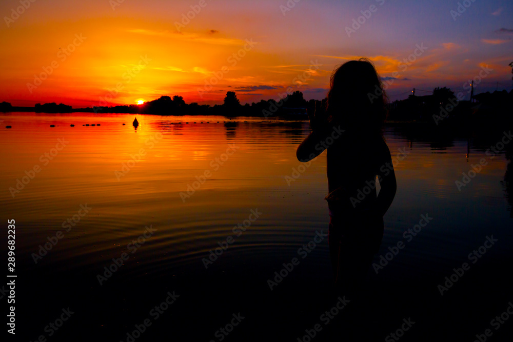 Fototapeta premium Silhouette of child by the lake and the orange sky in sunset